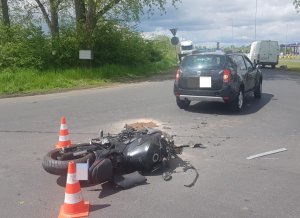Na zdjęciu widoczny jest motocykl leżący na boku, obok stojące czerwone słupki drogowe. Dalej widać samochód osobowy stojący tyłem.